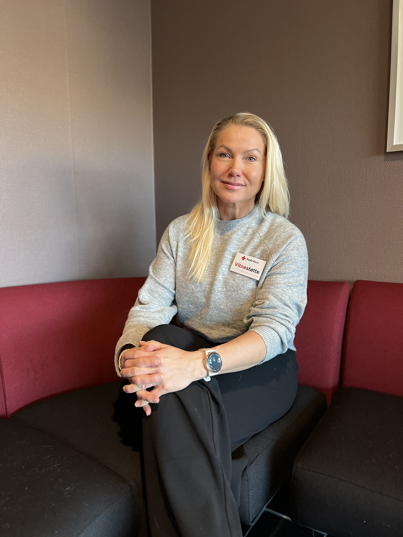 Kvinne sitter i en svart og rød hjørnesofa. Hun har langt lyst hår, lysegrå genser og en mørk genser.