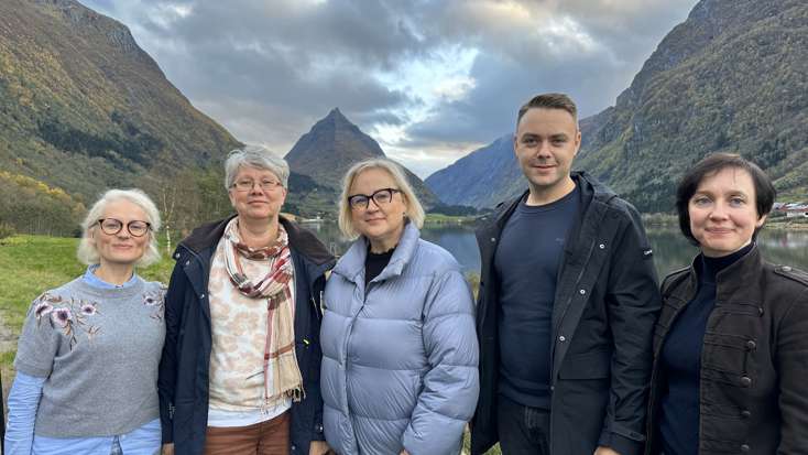 Fem smilende personer, fire kvinner og en mann, poserer foran vakker natur, med en spiss fjelltopp i bakgrunnen og med fjell og fjord på hver sin side i bildet, under lettskyet himmel..