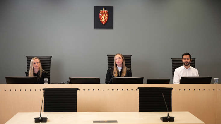 Fagommer og to meddommere sitter på hver sin stol bak dommerbord. På veggen bak henger riksvåpenet.