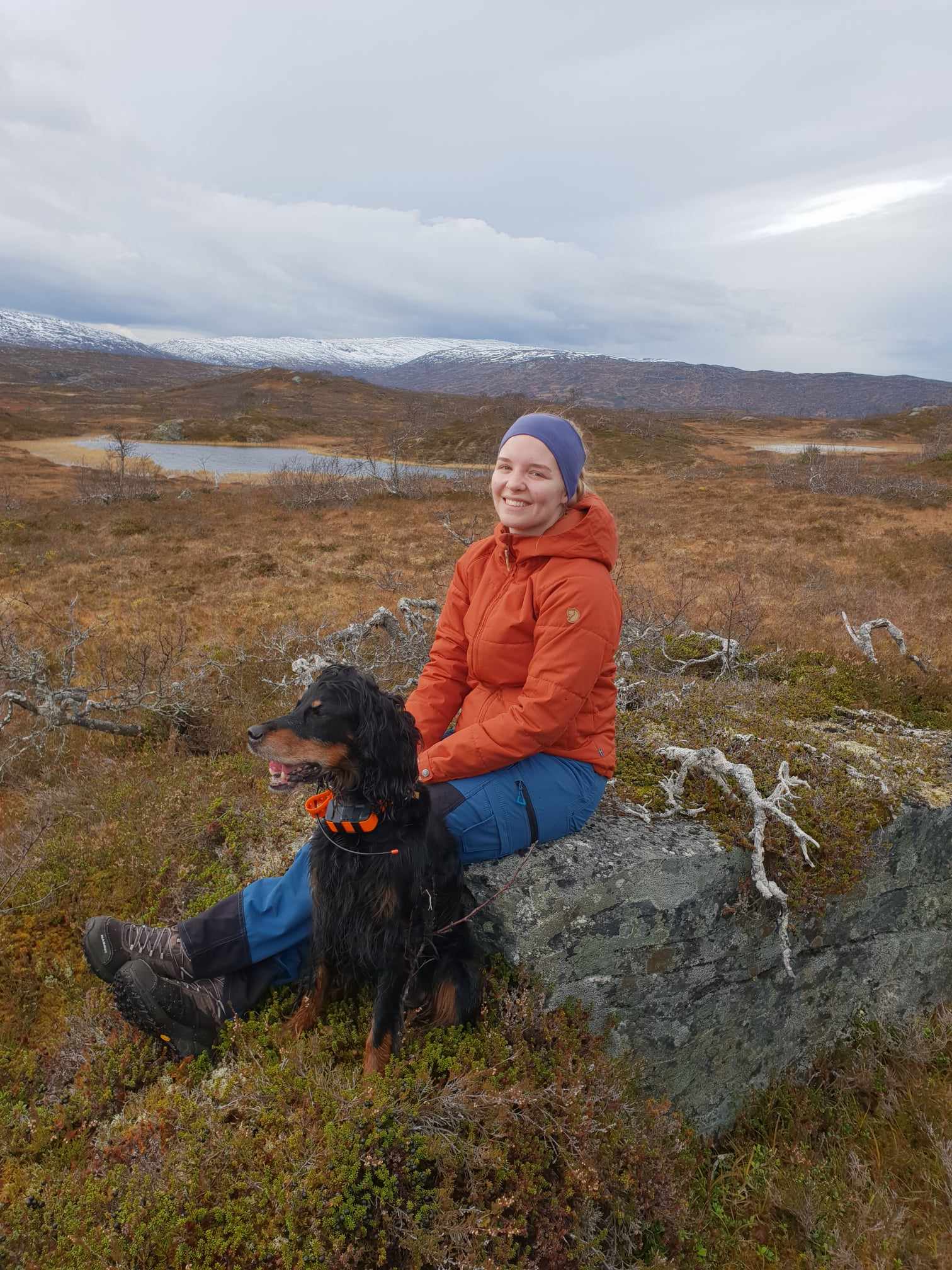 Kvinne sitter på en stor stein ute i myrlandskap. Hun har på seg oransje allværsjakke og blå turbukse, og et blått pannebånd. Det står en stor svart hund ved siden av seg.